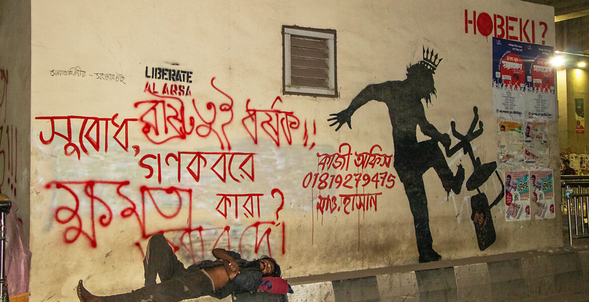 En hemlös man sover vid en vägg intill en väg, där någon skrivit: “Vad är i massgravarna, folket eller makten?” (översatt från bengali).