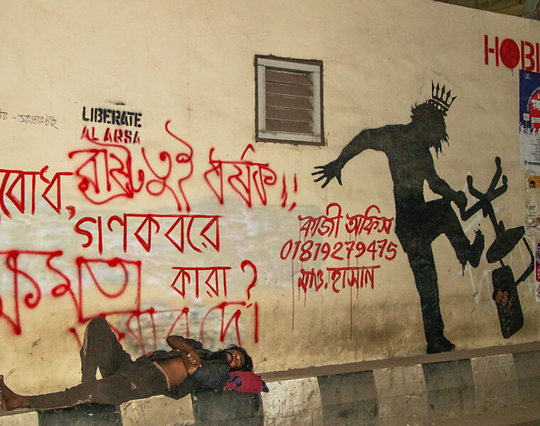 En hemlös man sover vid en vägg intill en väg, där någon skrivit: “Vad är i massgravarna, folket eller makten?” (översatt från bengali).